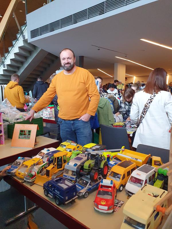 ein Mann in orangenem Pullover steht hinter seinem Verkaufstisch gefüllt mit Spielzeugautos und lächelt in die Kamera