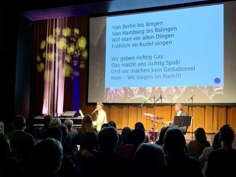 Am unteren Bildrand das Publikum, auf der Bühne Tobias Sudhoff am Klavier, neben ihm Gereon Homann am Schlagzeug, im Hintergrund eine Leinwand, die die Lyriks zeigt.