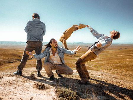 Drei Männer stehen in verschiedenen Posen in einer kargen Wüstenlandschaft