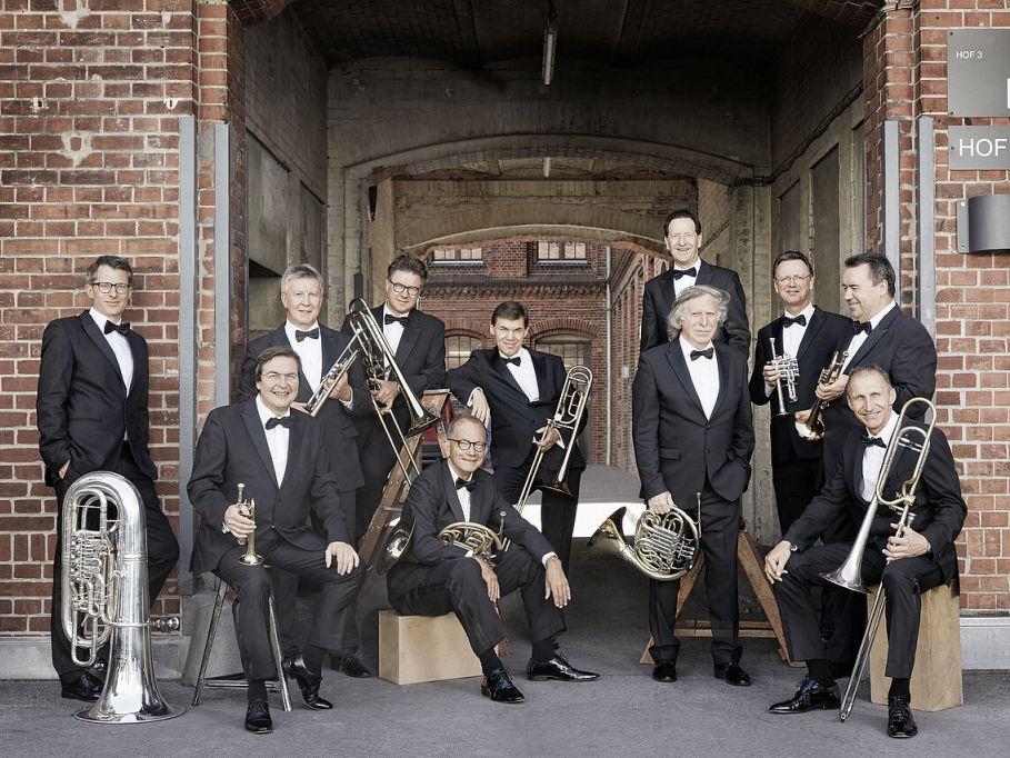 Die elf Musiker des German Brass Ensembles stehen und sitzen mit ihren Instrumenten und gekleidet in schwarzen Anzügen vor einem gemauerten Gebäude.