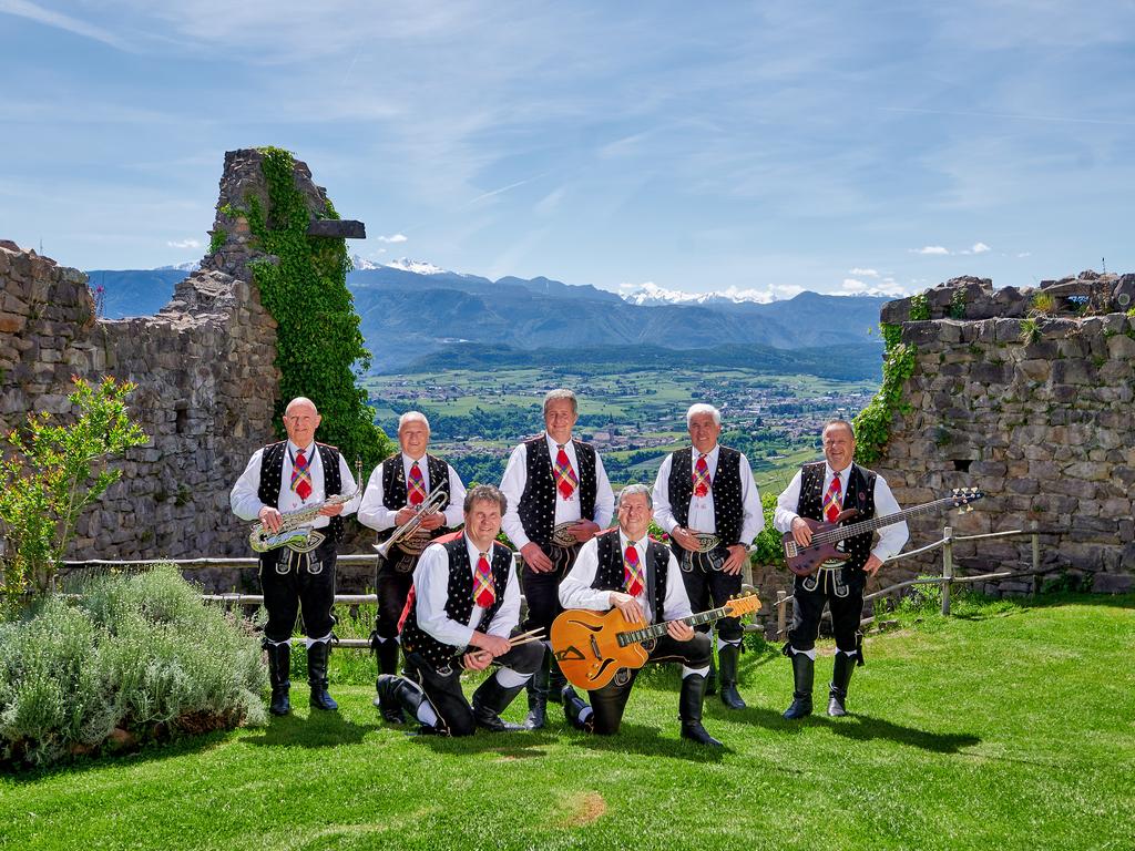 Bandfoto der sieben Musiker in traditioneller Tracht vor demHintergrund der Berge auf einer Burgruine