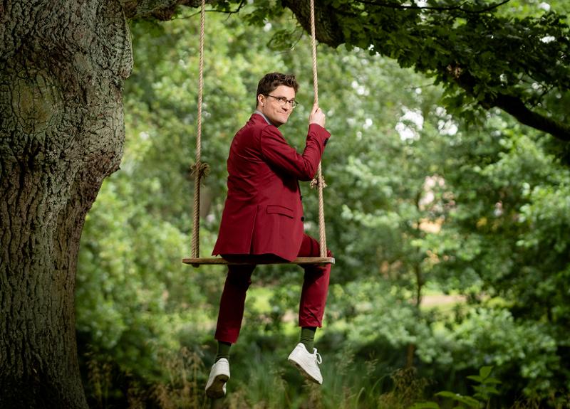 Bodo Wartke sitzt in rotem Anzug auf einer Schaukel, die an einem Baum hängt