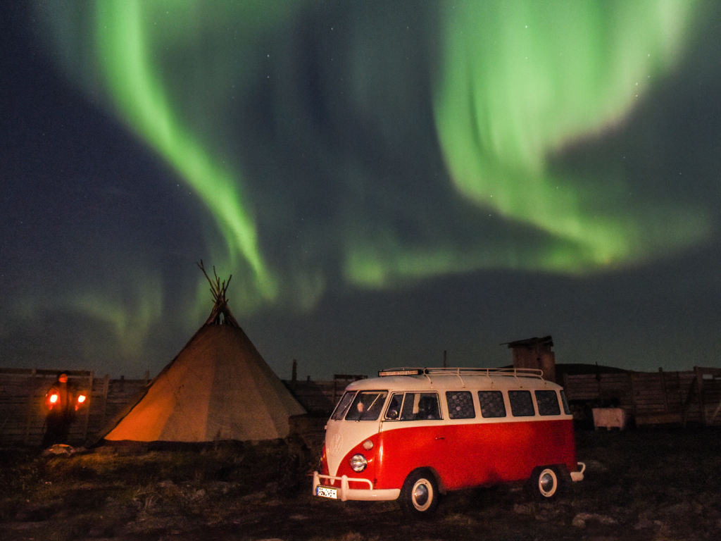 Der rote T1-Bulli "Erwin" steht im Vordergrund, links im Hintergrund ein Tippi. Am Nachthimmel grüne Nordlichter.